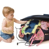 Baby girl kneeling near the suitcase, trying to pack it, isolated