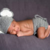 Thirteen day old smiling newborn baby boy wearing a gray crocheted elephant hat and diaper cover. He is sleeping on his stomach on gray fleece fabric.