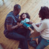 A photo of happy parents tickling son on hardwood floor. High angle view of family are enjoying in new house. They are in casuals. Young parents playing with child at home.