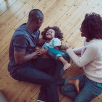A photo of happy parents tickling son on hardwood floor. High angle view of family are enjoying in new house. They are in casuals. Young parents playing with child at home.